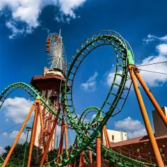Zielona kolejka górska z pomarańczowymi podporami, w tle wieża widokowa i diabelski młyn. Ten tilt coaster zapowiada emocjonującą przejażdżkę.