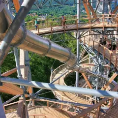Drewniana konstrukcja z metalową zjeżdżalnią tworzy ścieżkę w koronach drzew. Ludzie spacerują po platformach, podziwiając widoki. To wspaniały sky walk!