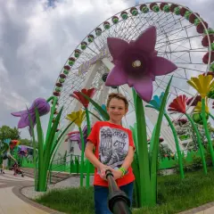 Chłopiec pozuje do selfie na tle diabelskiego młyna w chorzowskim wesołym miasteczku, otoczony kolorowymi, gigantycznymi kwiatami.