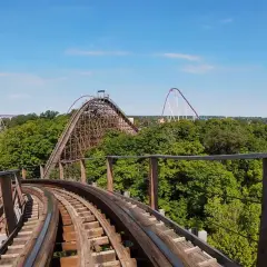 Widok z kolejki górskiej na tle zielonych drzew i błękitnego nieba. W oddali widać inne atrakcje parku rozrywki, w tym największy drewniany rollercoaster na świecie.