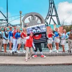 Ekipa w strojach filmowych pozuje przed logo Movie Park Germany. Wesoła ekipa z maskotką klapsa.
