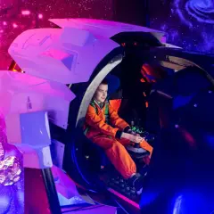 Chłopiec w pomarańczowym skafandrze steruje statkiem kosmicznym w Kosmoparku Wrocław, otoczony asteroidami i mgławicami.