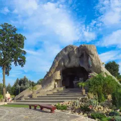 Jaskinia w skale, jedna z lubliniec atrakcji, z figurami i schodami, otoczona zielenią pod błękitnym niebem.