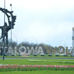 Pomnik i napis "STALOWA WOLA" w parku, otoczone kwitnącymi tulipanami.