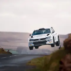Biały samochód rajdowy zespołu Energylandia Rally Team w locie nad wzniesieniem. Widzowie filmują akcję.