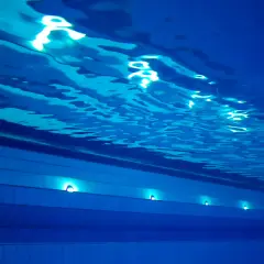Podwodne ujęcie basenu sportowego na Termach Maltańskich. Błękitna woda, falująca powierzchnia i światła podwodne tworzą spokojną atmosferę.