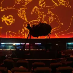 Wnętrze planetarium Warszawa. Na pomarańczowej kopule widnieją złote rysunki gwiazdozbiorów. Na środku stoi projektor.