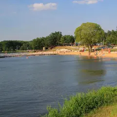 Słoneczny dzień nad jeziorem w Siemiatyczach. Plaża pełna ludzi, pomost z altaną, a w tle las i budynki. Idealne siemiatycze atrakcje.