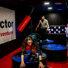 Kobieta pokonuje przeszkody w **blue city park rozrywki**. Mężczyźni obserwują jej zmagania.