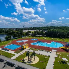 Letni dzień nad wodą. Widać rozległy kompleks basenów, zjeżdżalnie i ludzi odpoczywających na trawie.