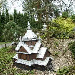 Drewniana makieta cerkwi z metalowym dachem, część parku miniatur centrum kultury ekumenicznej, otoczona zielenią i kamieniami.