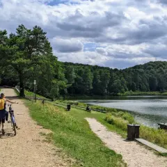 Kobieta z rowerem na ścieżce nad jeziorem, otoczonym lasem. W oddali widać wzgórza, a na wodzie pływają kaczki. To idealne miejsce na spacer, jak złoty potok.