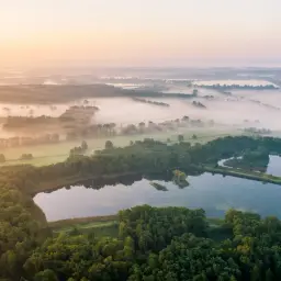 Mglisty poranek nad stawami w Dolinie Baryczy. Widok z lotu ptaka na lasy, pola i wodę, ukazujący piękno przyrody i liczne atrakcje tego regionu.