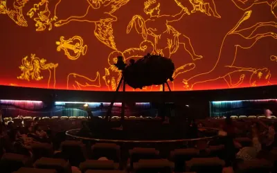 Wnętrze planetarium Warszawa. Na pomarańczowej kopule widnieją złote rysunki gwiazdozbiorów. Na środku stoi projektor.