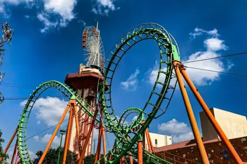 Zielona kolejka górska z pomarańczowymi podporami, w tle wieża widokowa i diabelski młyn. Ten tilt coaster zapowiada emocjonującą przejażdżkę.