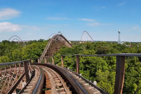 Widok z kolejki górskiej na tle zielonych drzew i błękitnego nieba. W oddali widać inne atrakcje parku rozrywki, w tym największy drewniany rollercoaster na świecie.