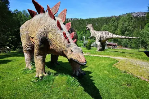 W parku dinozaurów karłów Stegozaur pochyla się do trawy, podczas gdy Tyranozaur czuwa w oddali.