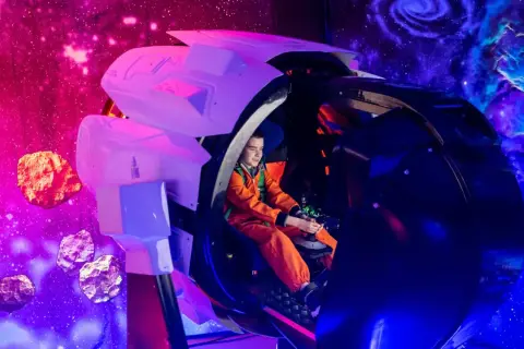 Chłopiec w pomarańczowym skafandrze steruje statkiem kosmicznym w Kosmoparku Wrocław, otoczony asteroidami i mgławicami.