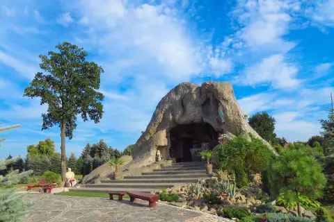 Jaskinia w skale, jedna z lubliniec atrakcji, z figurami i schodami, otoczona zielenią pod błękitnym niebem.