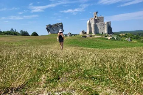 Kobieta w kapeluszu idzie przez łąkę w kierunku ruin zamku na Szlaku Orlich Gniazd.