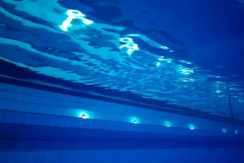 Podwodne ujęcie basenu sportowego na Termach Maltańskich. Błękitna woda, falująca powierzchnia i światła podwodne tworzą spokojną atmosferę.