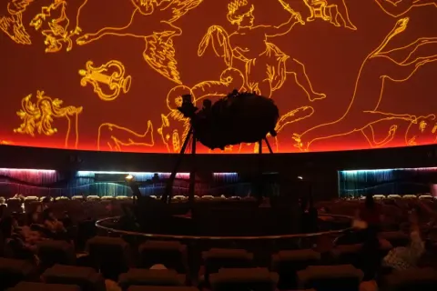 Wnętrze planetarium Warszawa. Na pomarańczowej kopule widnieją złote rysunki gwiazdozbiorów. Na środku stoi projektor.