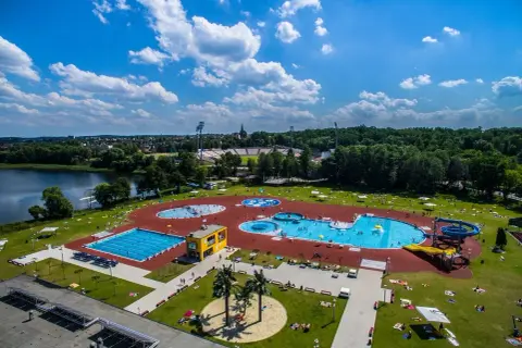 Letni dzień nad wodą. Widać rozległy kompleks basenów, zjeżdżalnie i ludzi odpoczywających na trawie.