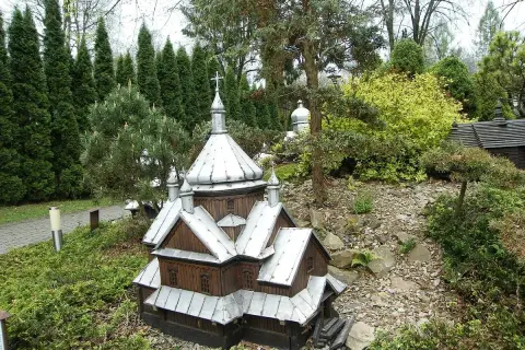 Drewniana makieta cerkwi z metalowym dachem, część parku miniatur centrum kultury ekumenicznej, otoczona zielenią i kamieniami.
