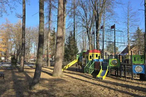 Kolorowy plac zabaw w parku rozrywki Nowy Sącz, z drabinkami, zjeżdżalniami i siatką do wspinaczki, otoczony drzewami.