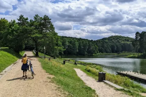 Kobieta z rowerem na ścieżce nad jeziorem, otoczonym lasem. W oddali widać wzgórza, a na wodzie pływają kaczki. To idealne miejsce na spacer, jak złoty potok.