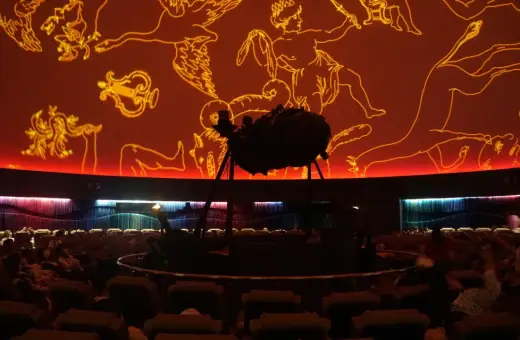 Wnętrze planetarium Warszawa. Na pomarańczowej kopule widnieją złote rysunki gwiazdozbiorów. Na środku stoi projektor.