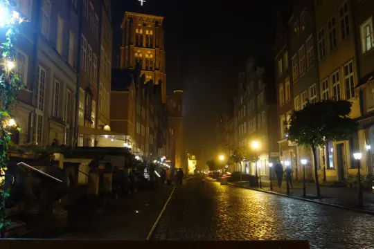 Mroczny, brukowany zaułek w Gdańsku nocą. Latarnie rzucają złote światło na mokrą nawierzchnię, a w oddali widać wieżę kościoła.
