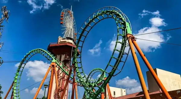 Zielona kolejka górska z pomarańczowymi podporami, w tle wieża widokowa i diabelski młyn. Ten tilt coaster zapowiada emocjonującą przejażdżkę.