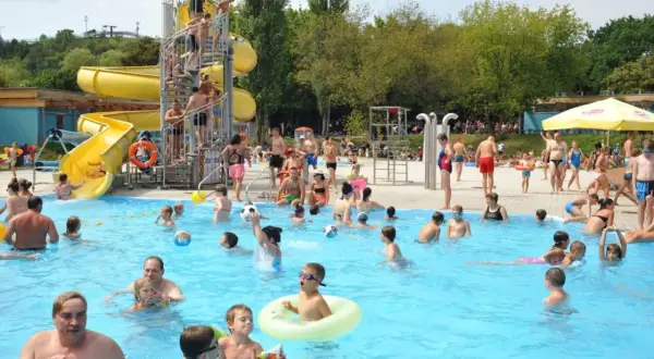 Radosny tłum ludzi korzysta z atrakcji basenu odkrytego w Warszawie. Dzieci bawią się w wodzie, zjeżdżają ze zjeżdżalni i grają w piłkę.