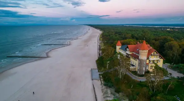 Zachód słońca nad jeziorem Łebsko i plażą. W oddali widać zalesione wybrzeże i malowniczy budynek z wieżą.
