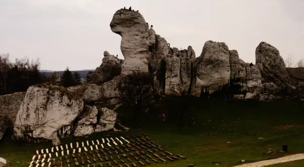 Skalne miasto podzamcza z turystami na szczytach. W dole rzędy drewnianych ławek na trawiastym placu.