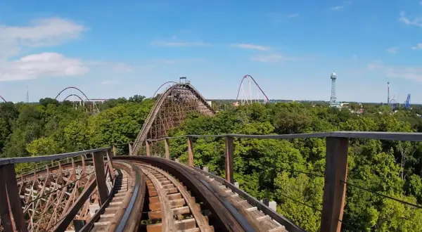 Widok z kolejki górskiej na tle zielonych drzew i błękitnego nieba. W oddali widać inne atrakcje parku rozrywki, w tym największy drewniany rollercoaster na świecie.