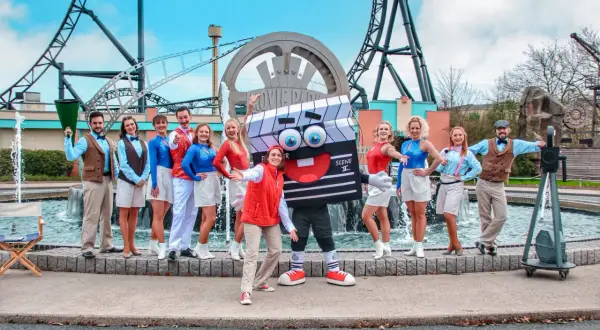 Ekipa w strojach filmowych pozuje przed logo Movie Park Germany. Wesoła ekipa z maskotką klapsa.
