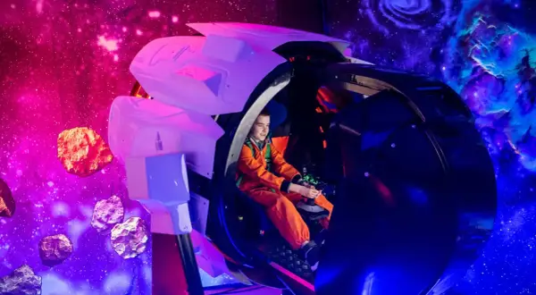 Chłopiec w pomarańczowym skafandrze steruje statkiem kosmicznym w Kosmoparku Wrocław, otoczony asteroidami i mgławicami.