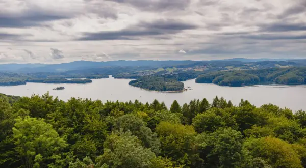 Widok na jezioro solińskie otoczone zielonymi wzgórzami i lasami pod pochmurnym niebem.