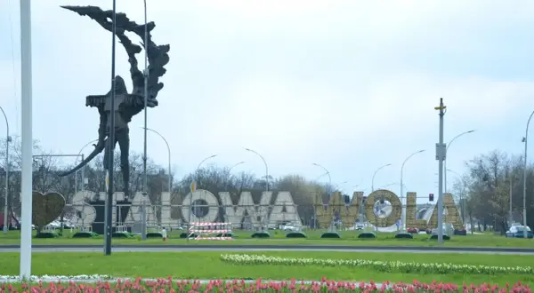 Pomnik i napis "STALOWA WOLA" w parku, otoczone kwitnącymi tulipanami.