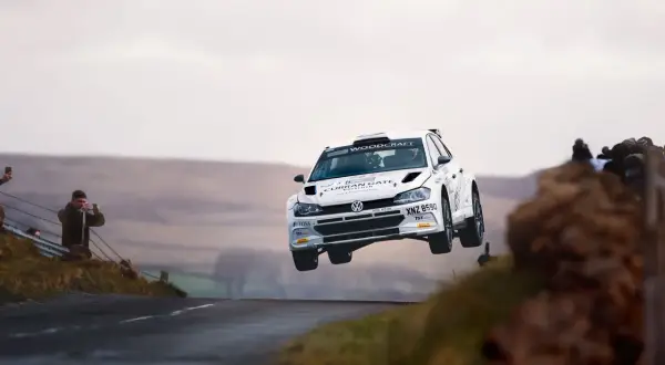 Biały samochód rajdowy zespołu Energylandia Rally Team w locie nad wzniesieniem. Widzowie filmują akcję.