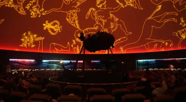 Wnętrze planetarium Warszawa. Na pomarańczowej kopule widnieją złote rysunki gwiazdozbiorów. Na środku stoi projektor.