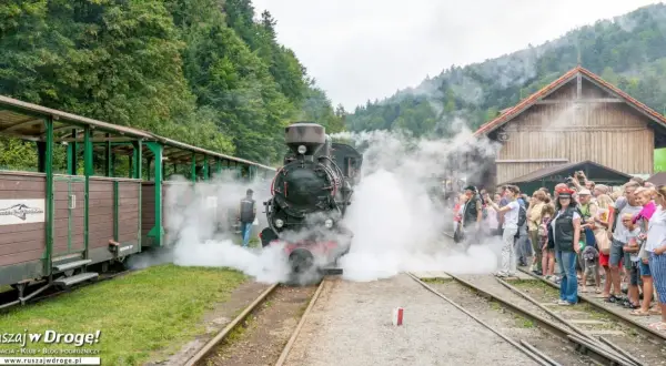 Parowóz z wagonami na tle górskich lasów. Tłum ludzi, w tym dzieci, obserwuje parę wydobywającą się z lokomotywy. Bieszczady atrakcje dla dzieci.