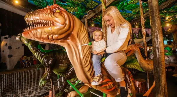 Uśmiechnięta mama z dzieckiem na dinozaurze w góraszka park rozrywki. Wesoła przejażdżka wśród prehistorycznych stworzeń.