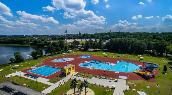 Letni dzień nad wodą. Widać rozległy kompleks basenów, zjeżdżalnie i ludzi odpoczywających na trawie.