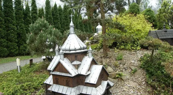 Drewniana makieta cerkwi z metalowym dachem, część parku miniatur centrum kultury ekumenicznej, otoczona zielenią i kamieniami.