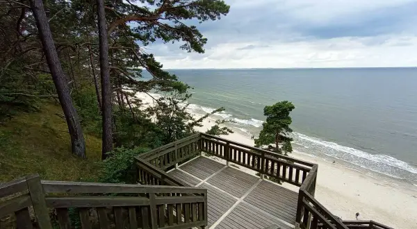 Drewniane schody prowadzą na plażę z widokiem na morze. W oddali widać las i **kaczą górę**.