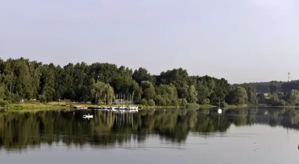 Spokojne jezioro Przeczyckie z odbijającymi się w wodzie drzewami i żaglówkami. Na wodzie widać samotną łódkę.