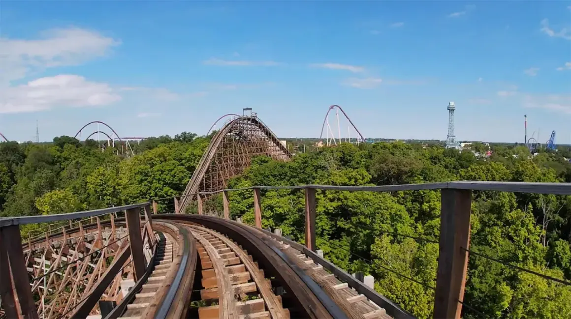 Widok z kolejki górskiej na tle zielonych drzew i błękitnego nieba. W oddali widać inne atrakcje parku rozrywki, w tym największy drewniany rollercoaster na świecie.